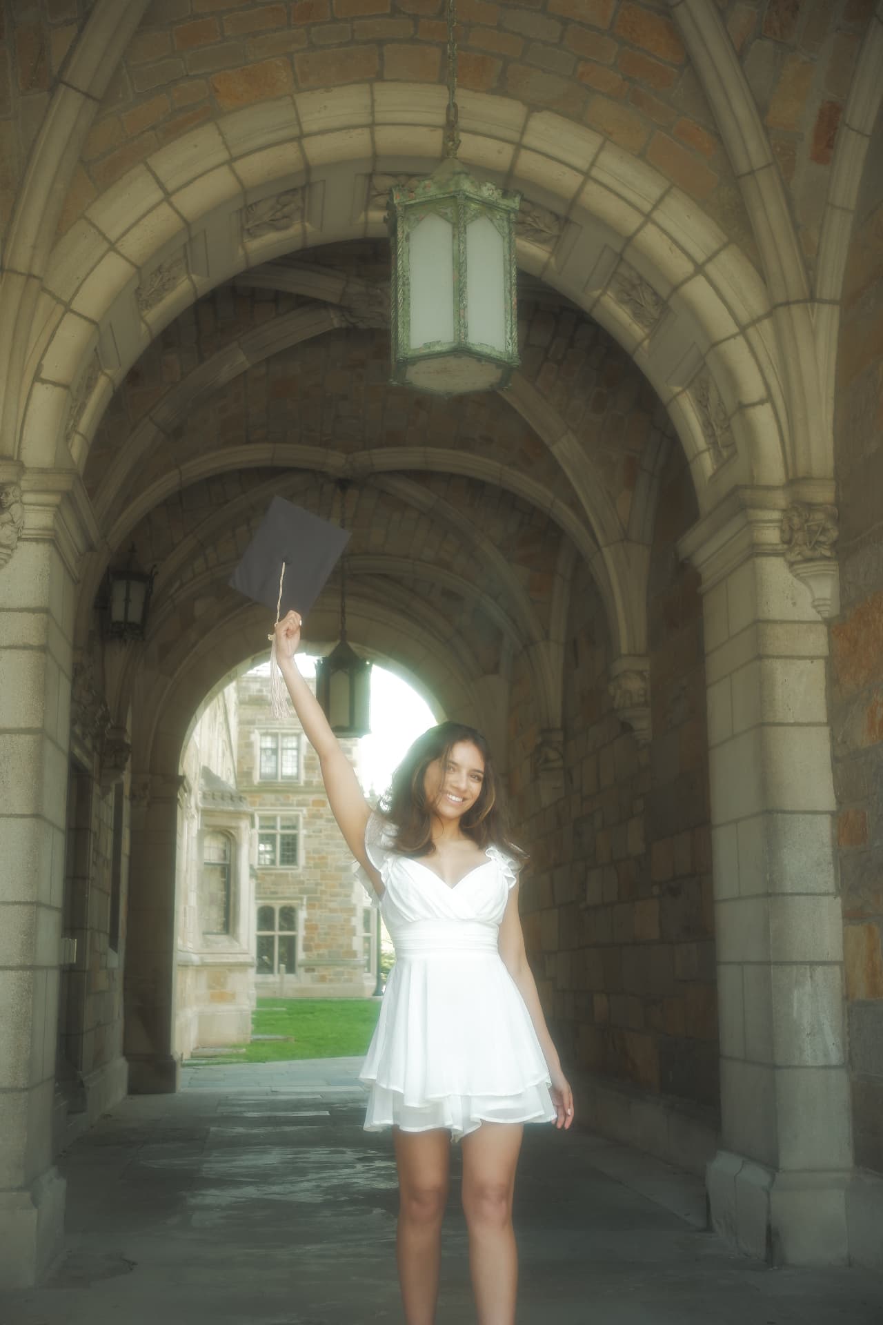Grace Varghese in a white dress holding a graduation cap under the University of Michigan Law Quad arches